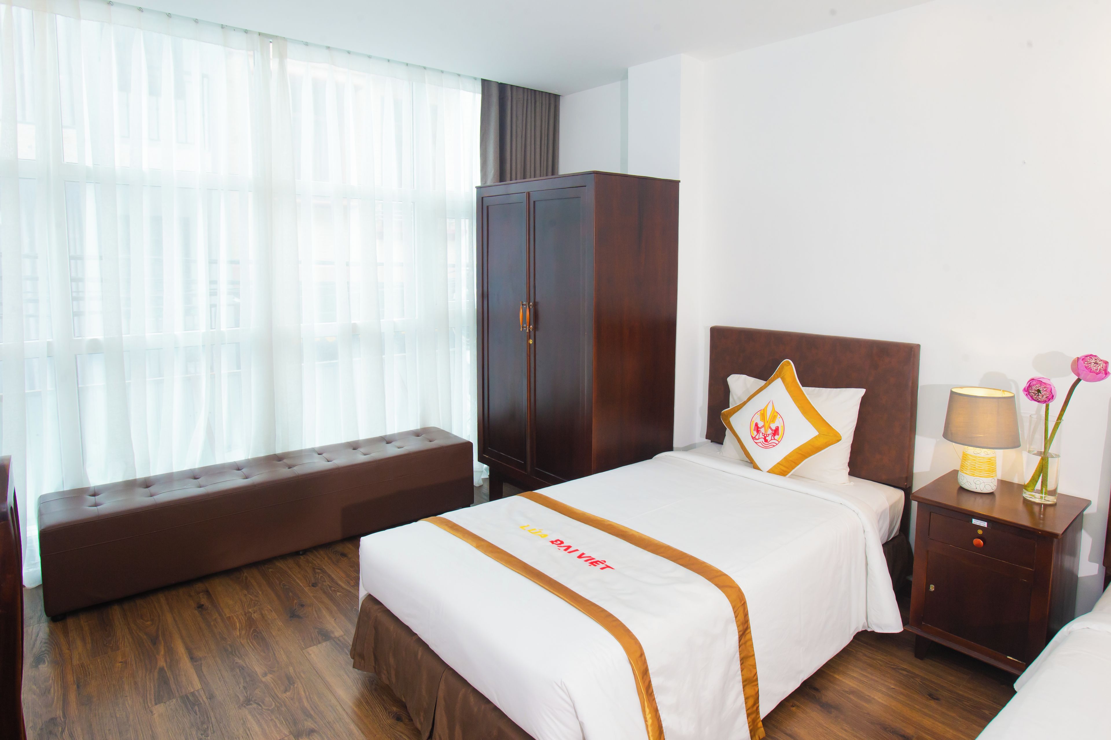 A hotel room with a bed, a bench, a wardrobe, a bedside table with a lamp, and a vase with flowers.