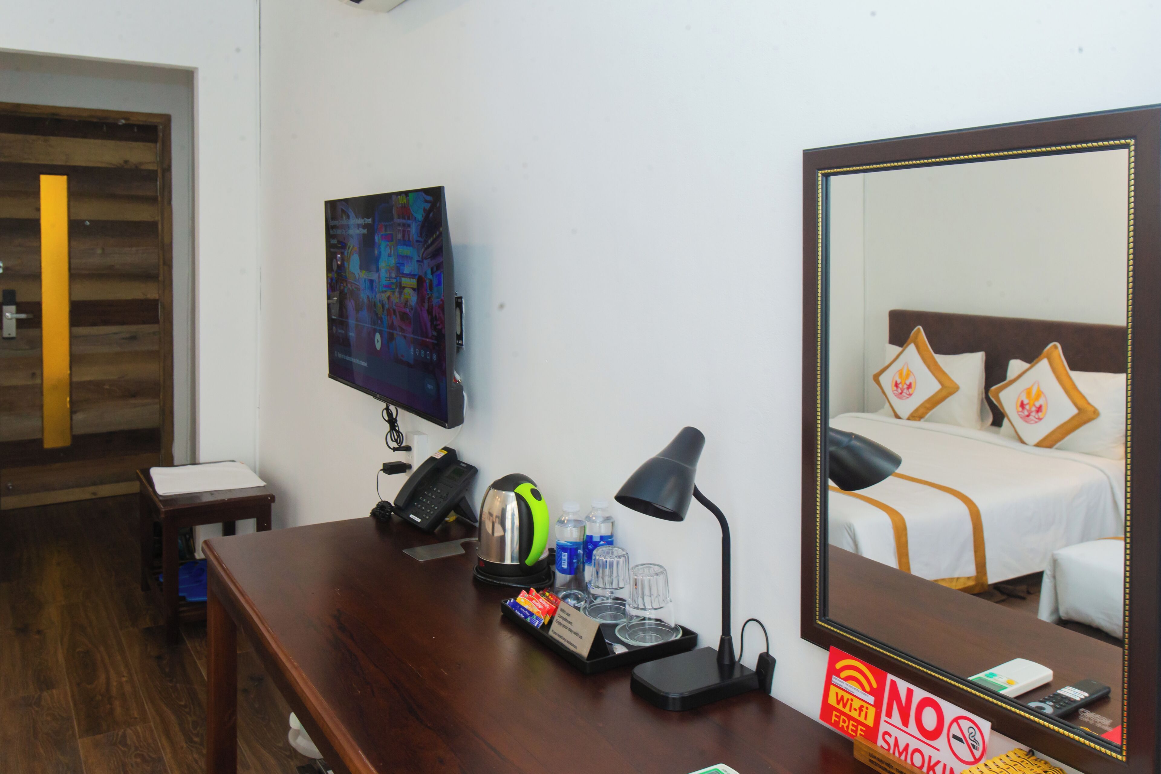 A hotel room with a wooden desk, a mirror, a bed with white bedding and decorative pillows, a television, and a 'No Smoking' sign.