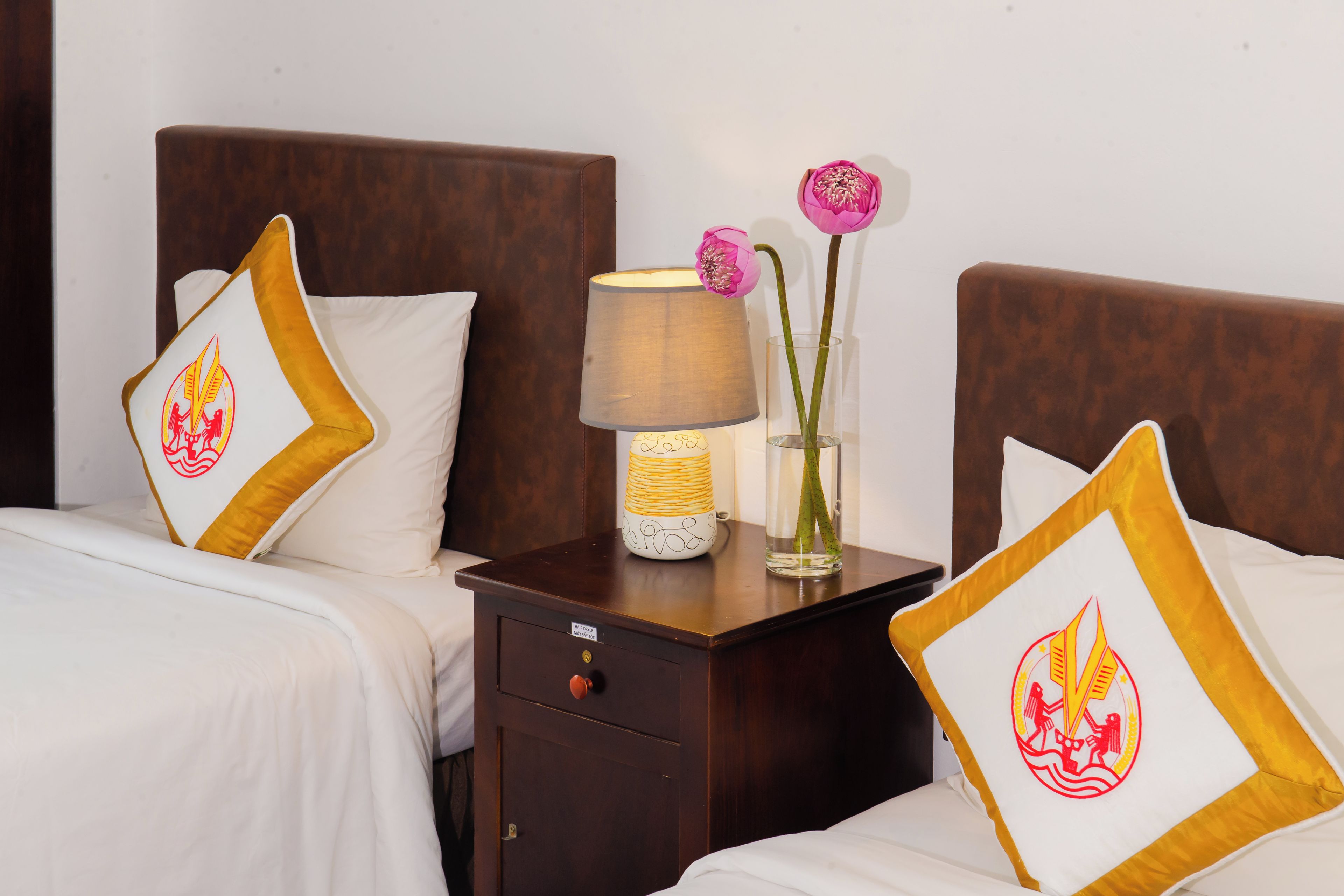 A hotel room with two beds, each with a pillow featuring a red and yellow emblem, a bedside table with a lamp and a vase with flowers, and a small drawer.