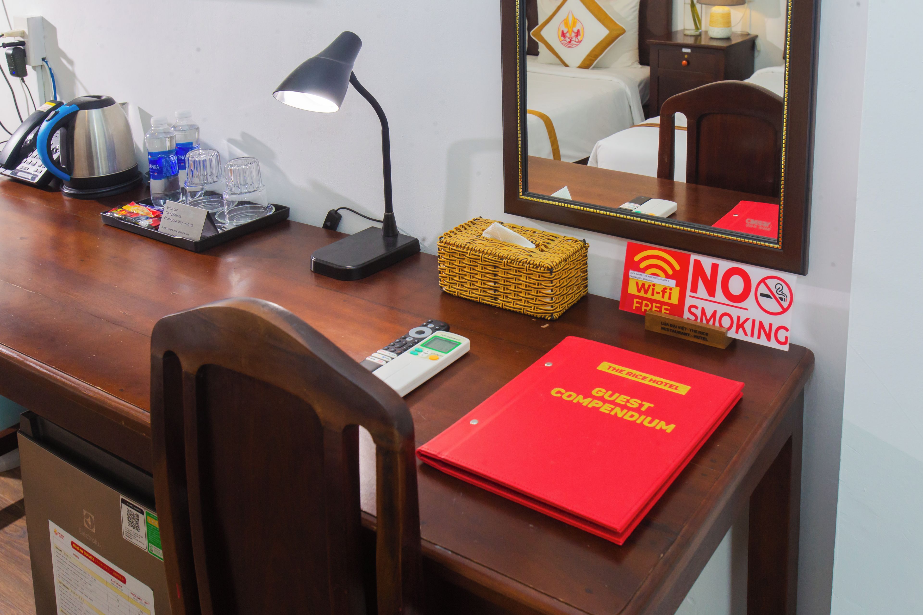 A hotel room desk with a laptop, a mobile phone, a remote control, a tray with glasses and a bottle, a red folder, and a 'NO SMOKING' sign.