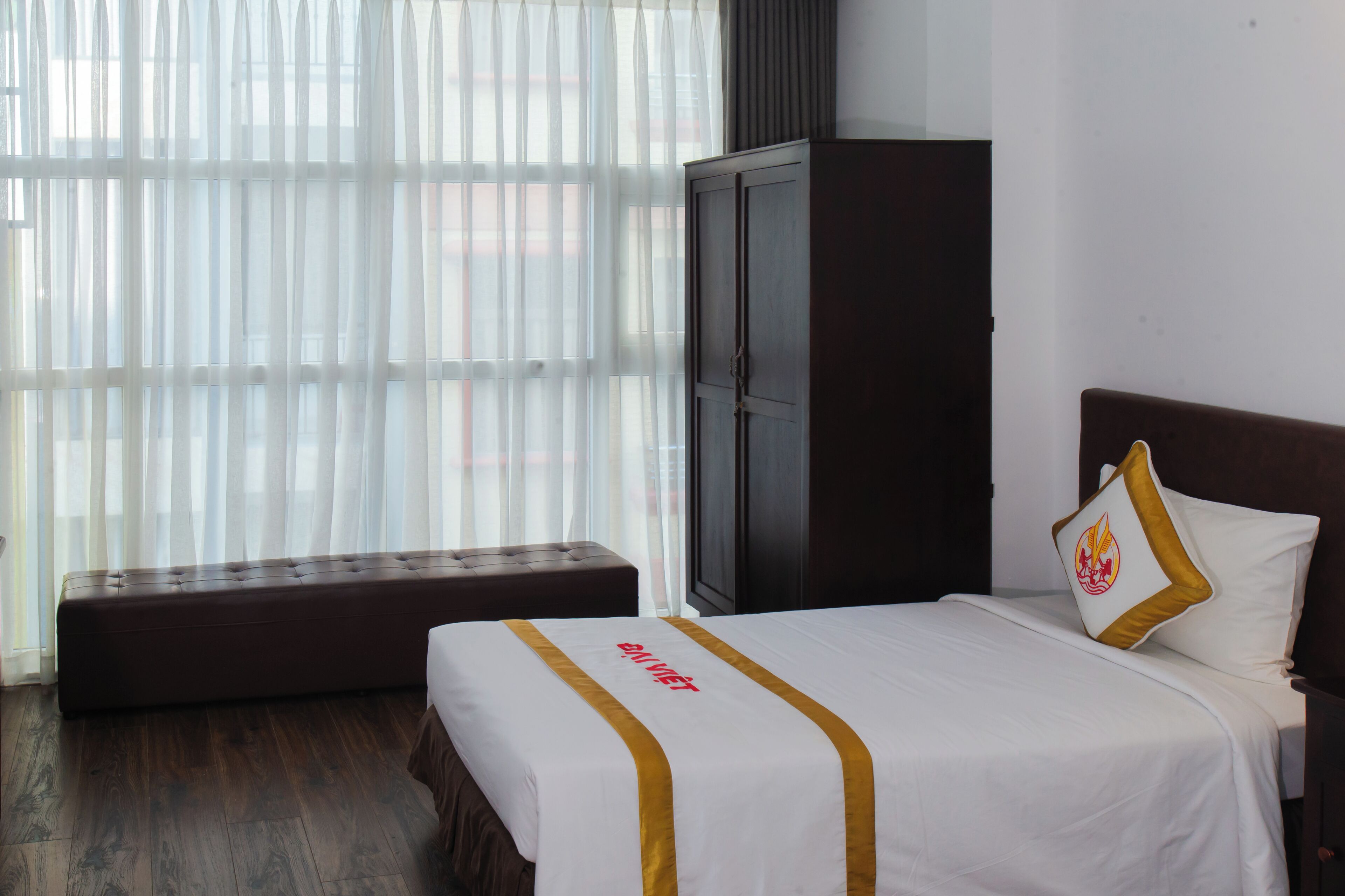 Una habitación de hotel con cama, un banco, un armario y una ventana con cortinas traslúcidas.