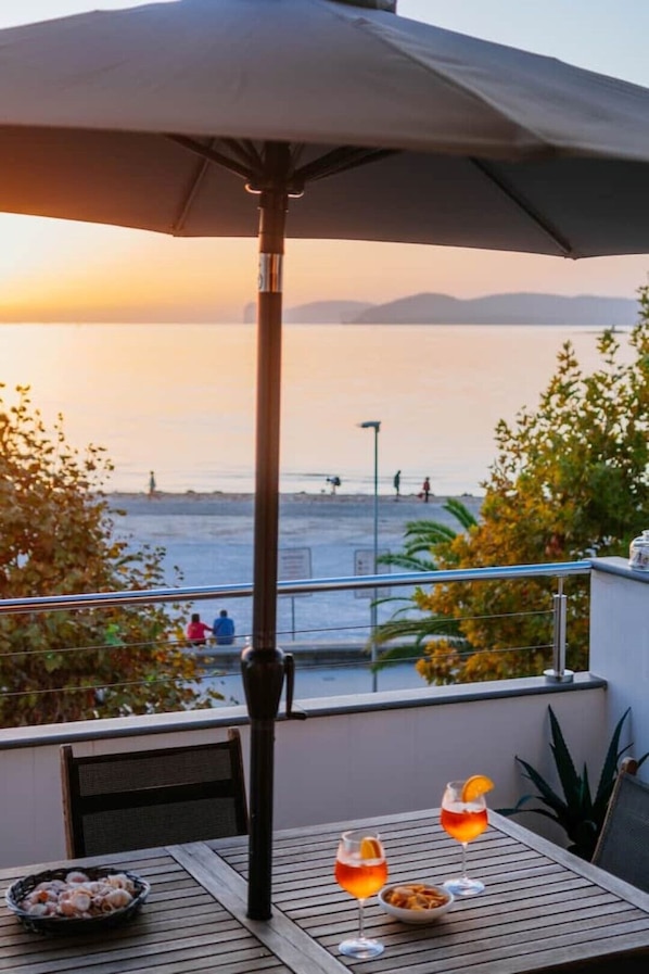 A balcony with a view of the sea at sunset, a table with drinks and snacks, and a large umbrella providing shade.