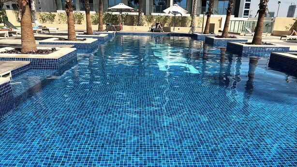 Una piscina con mosaicos azules, rodeada de sillas de descanso y palmeras.