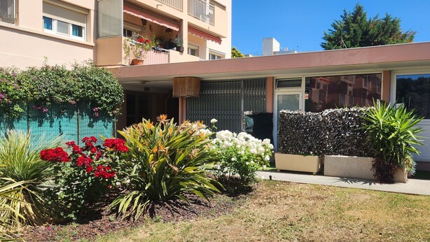 A modern building with a covered entrance, a balcony with potted plants, and a garden with various flowers and plants. - Logement Résidence (Antibes)