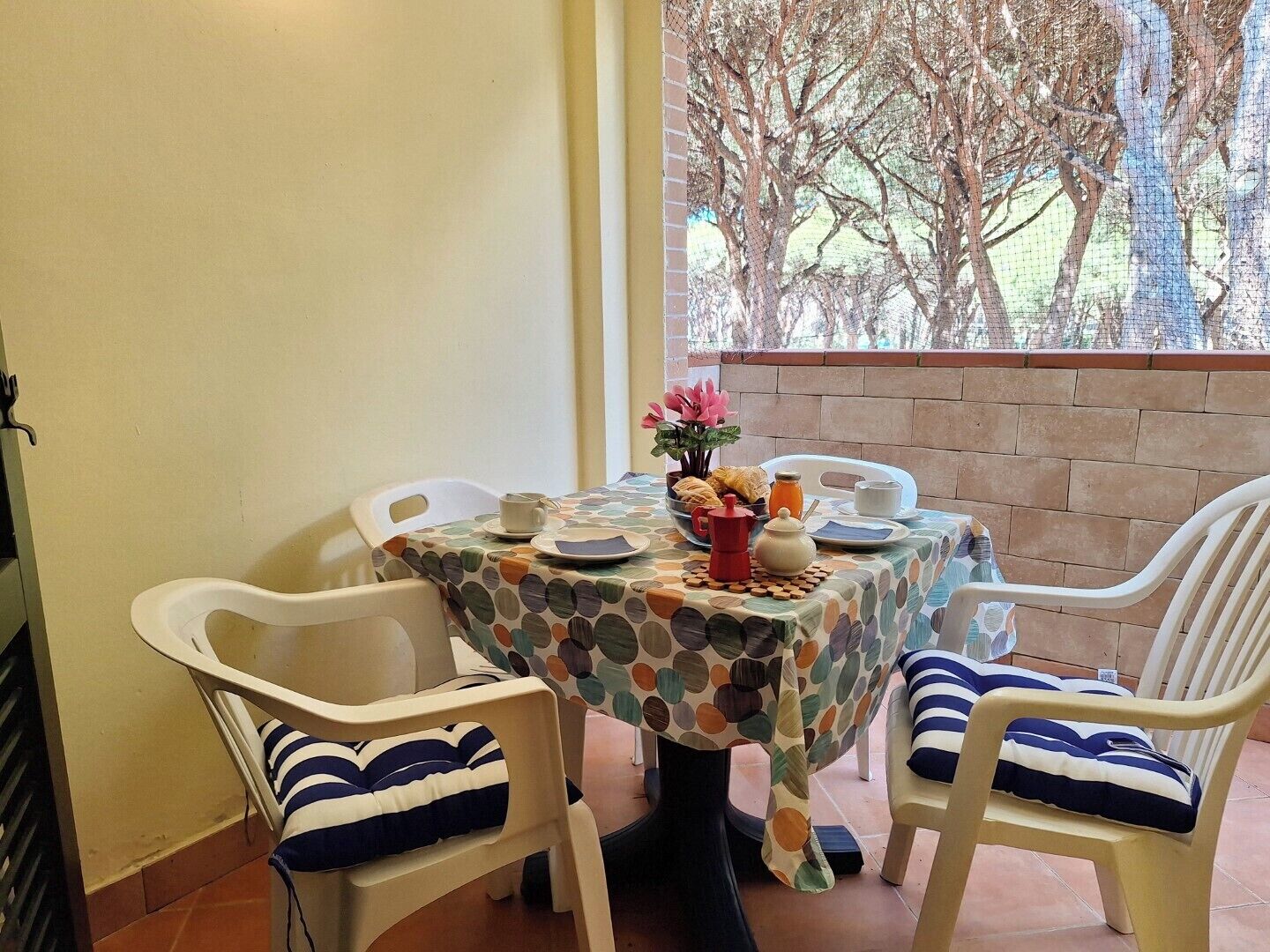 Un coin repas avec une table dressée pour deux personnes, agrémenté d’une nappe à pois, de chaises blanches avec des coussins à rayures et d’une vue sur les arbres par la fenêtre.