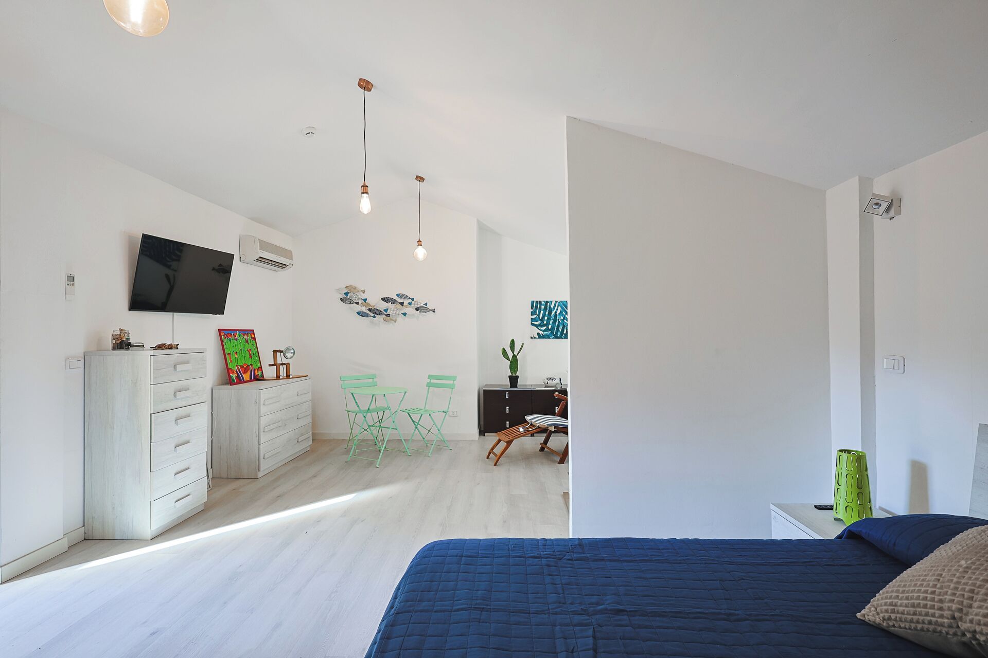 Un dormitorio moderno con una cama grande, un televisor montado en la pared y una zona de escritorio con una silla y una planta de cactus.
