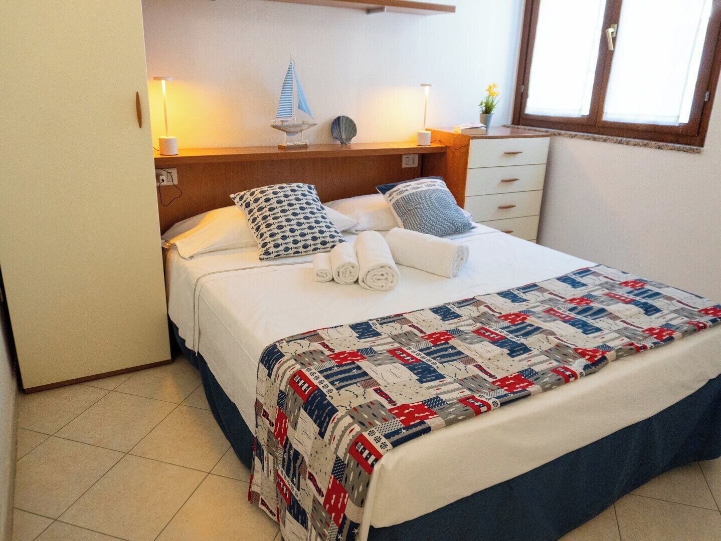 A neatly made bed with a quilt, pillows, and rolled towels. There are two bedside lamps and a small decorative item on the nightstand.