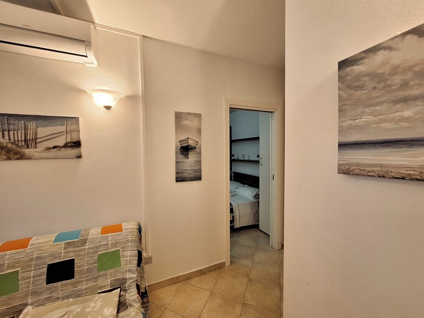 A hallway with a bed, a painting of a boat, and a painting of a beach.