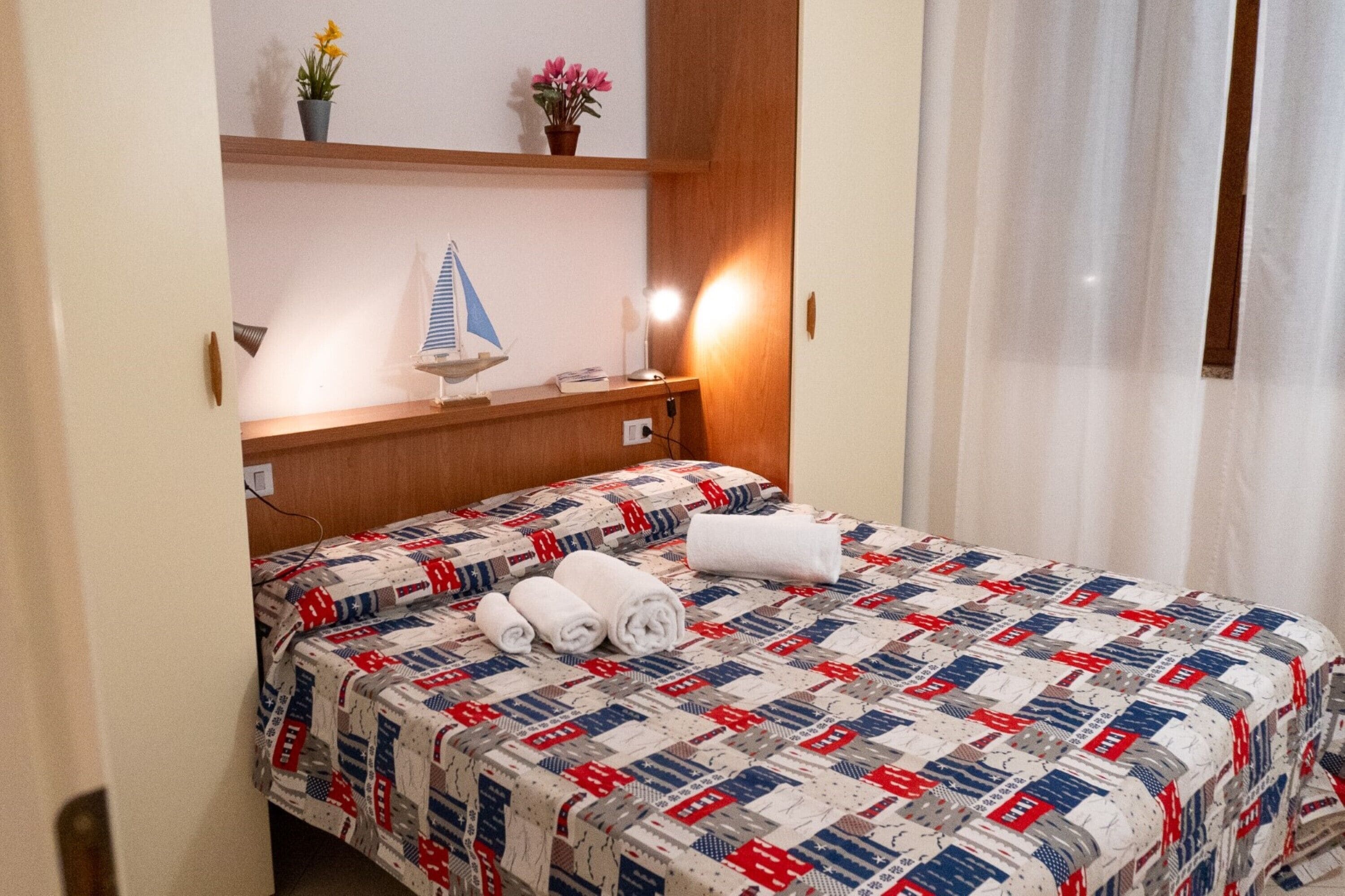 Una habitación de hotel con una cama, una mesita de noche, una lámpara y una estantería con objetos decorativos.