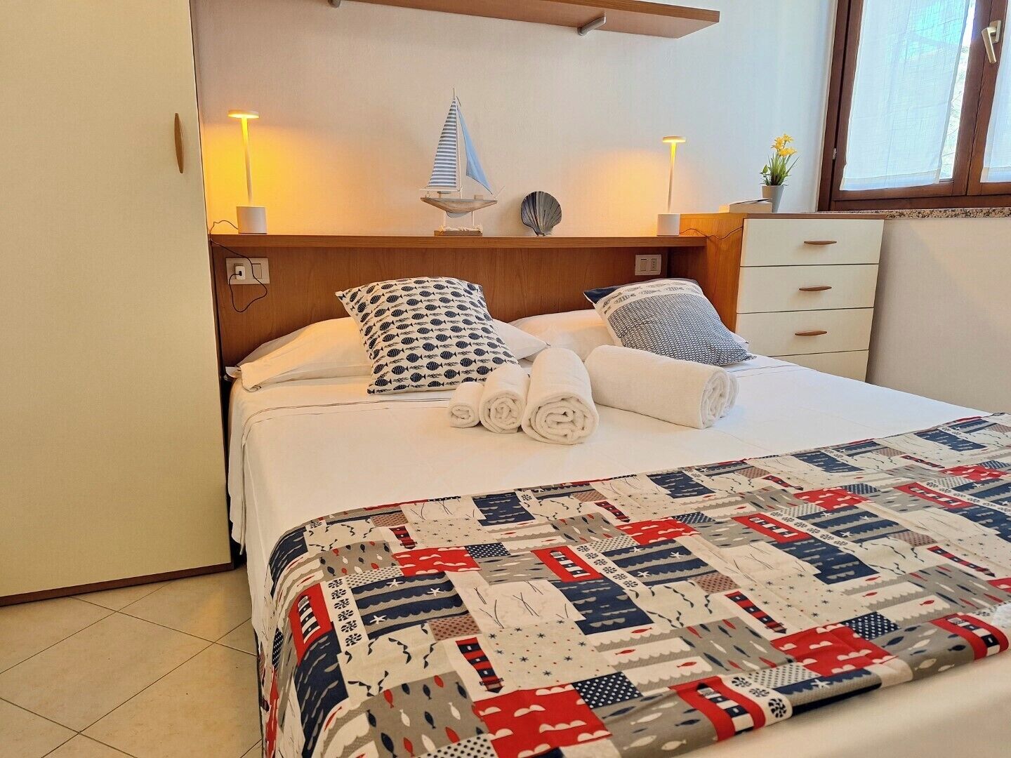 A neatly made bed with a quilt, pillows, and rolled towels. There is a small decorative sailboat on the headboard.