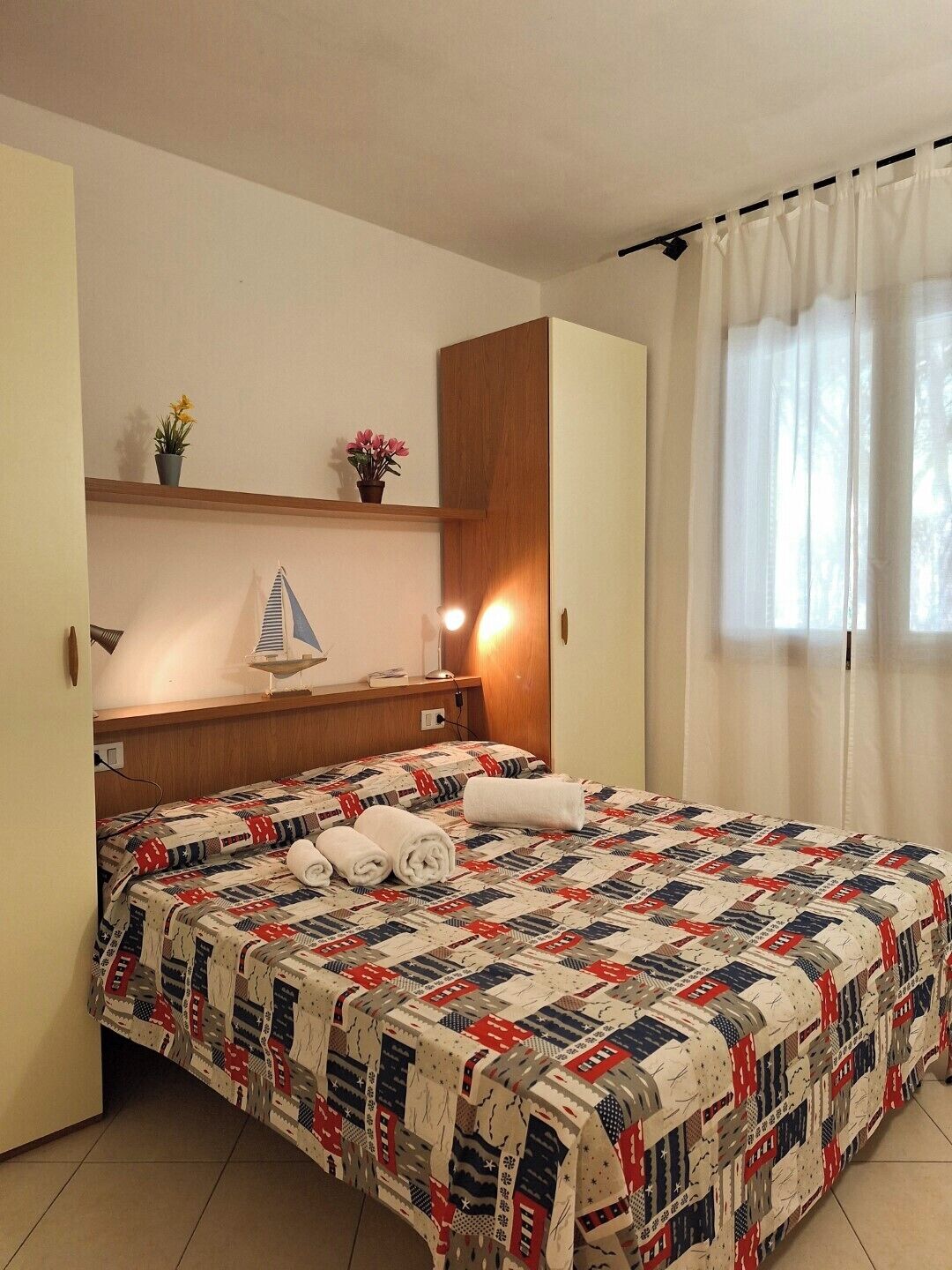 A hotel room with a bed, a nightstand, a shelf with decorative items, a window with curtains, and a wall-mounted lamp.