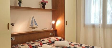 A hotel room with a bed, a nightstand, a shelf with decorative items, a window with curtains, and a wall-mounted lamp.