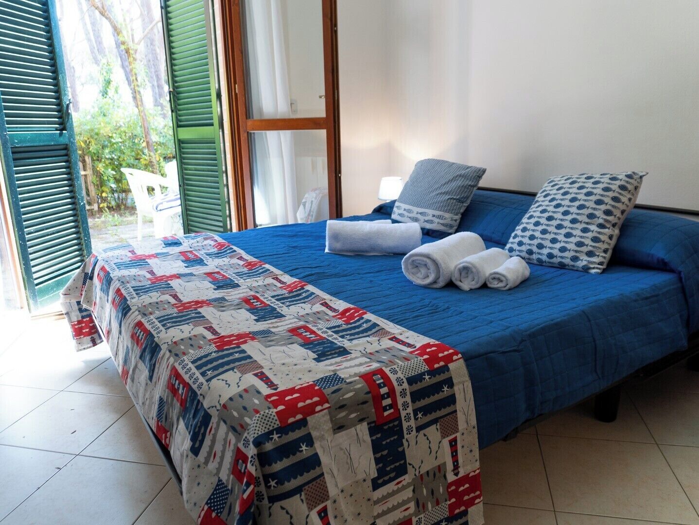 Een net opgemaakt bed met blauw bedlinnen en een sprei, kussens en opgerolde handdoeken op een betegelde vloer.