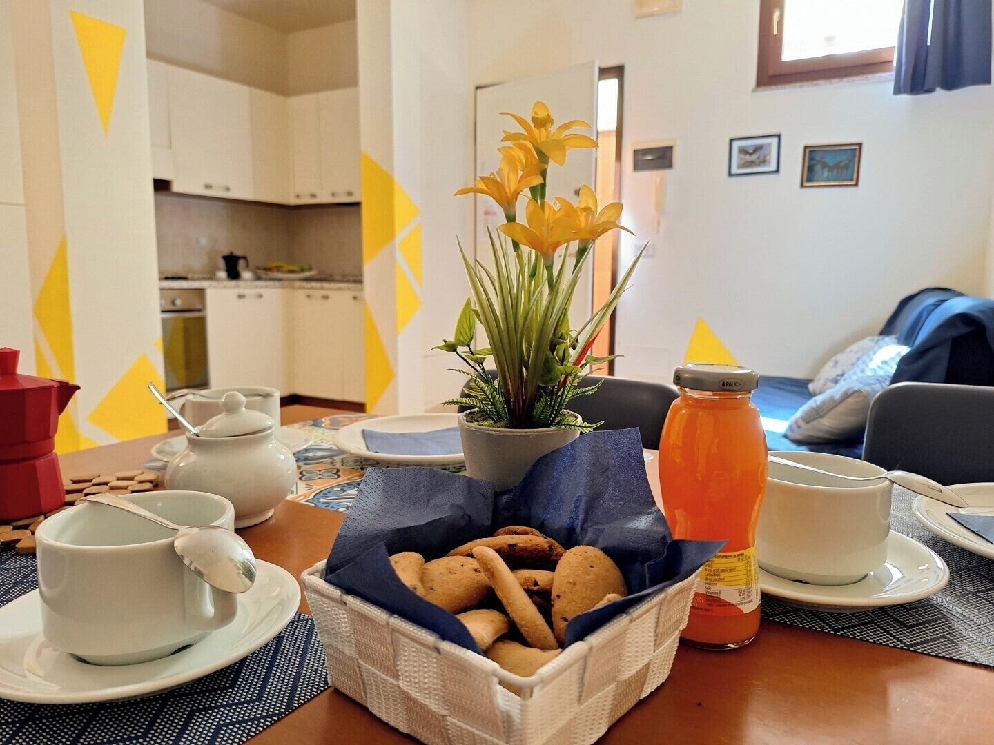 Una disposición para el desayuno con una taza de café, una tetera, un tarro de mermelada, una cesta de galletas y una planta en maceta.