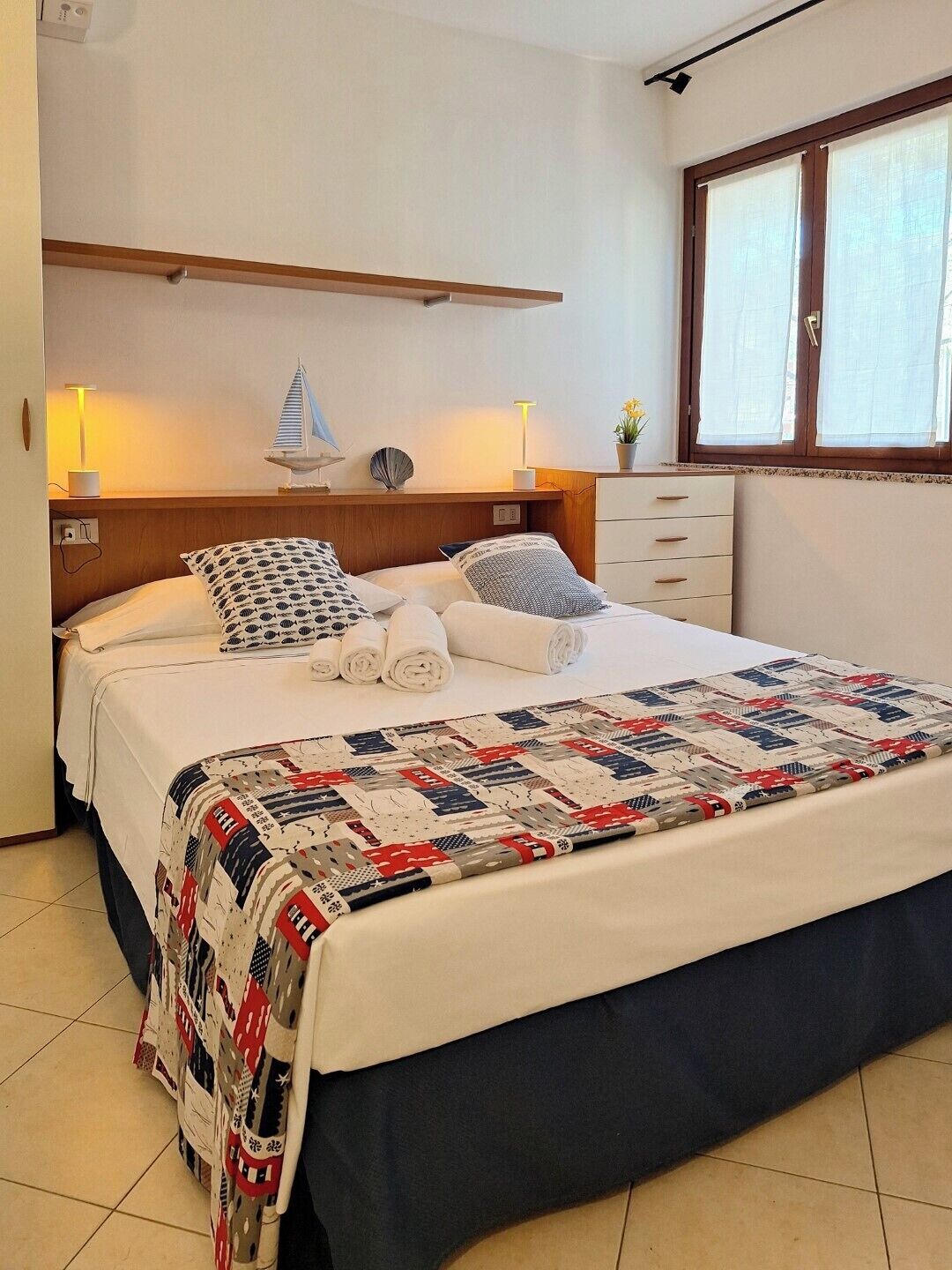 Una cama bien tendida con colcha, almohadas y toallas enrolladas. Se aprecia un pequeño estante con objetos decorativos y una lámpara.
