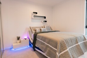 A modern bedroom with a bed, bedside tables, a wall-mounted shelf, and a small plant.