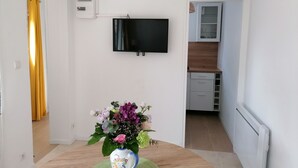 A modern living room with a flat-screen TV mounted on the wall, a decorative vase with flowers on a table, and a kitchen area visible through an open door.