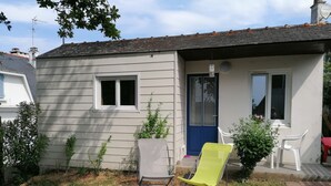 A small house with a paved patio, two windows, a blue door, and a small garden area with a chair and a table.