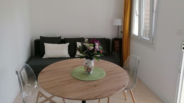 A dining area with a round wooden table, clear chairs, a vase with flowers, a dark sofa with cushions, a lamp, and a window with curtains.