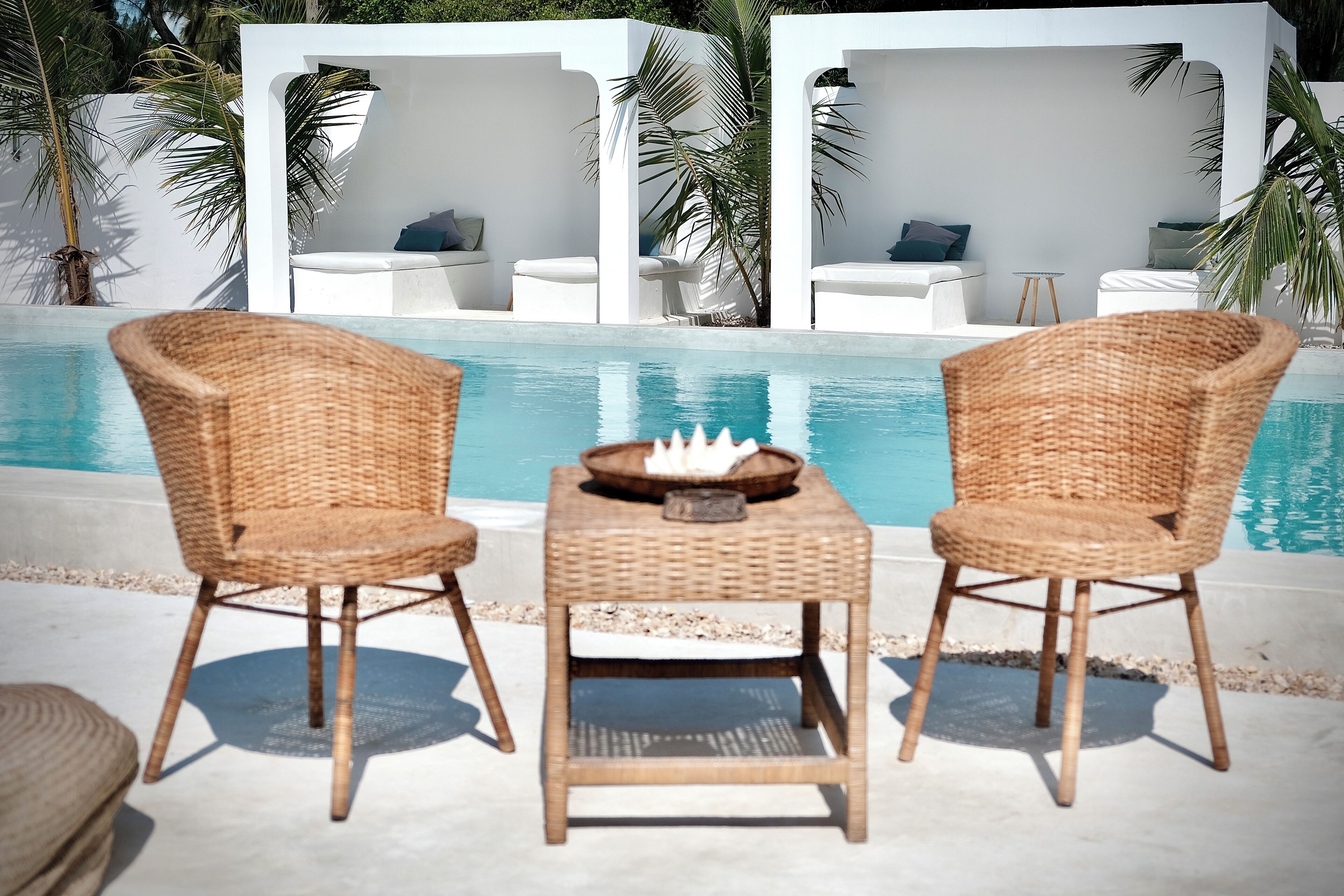 A poolside area with two wicker chairs, a small table with a bowl and a candle, and white lounge chairs with cushions.