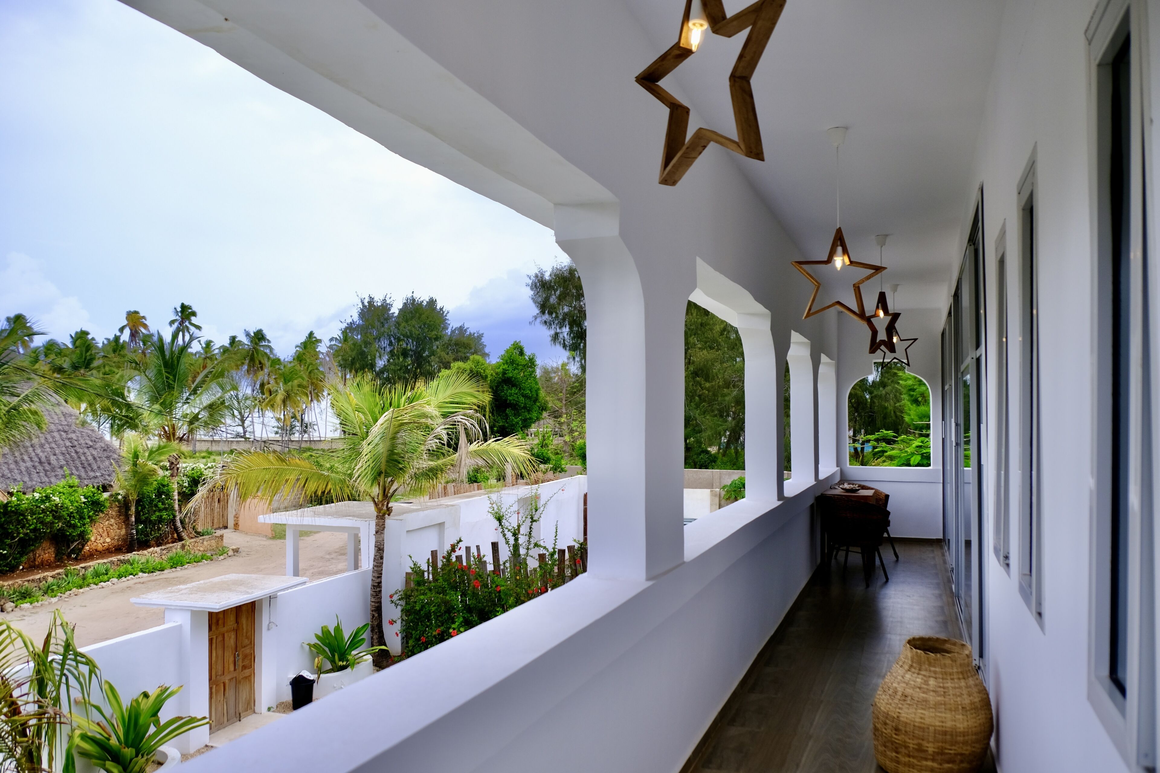 Un pasillo con paredes blancas, luces de techo en forma de estrella de madera y amplios ventanales que ofrecen vistas a un jardín exuberante y palmeras.