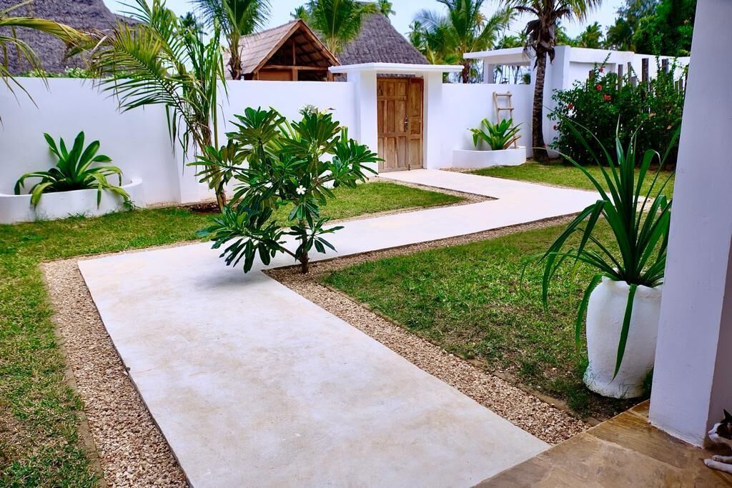 Uma área externa moderna com caminho de concreto, paredes brancas e plantas tropicais.