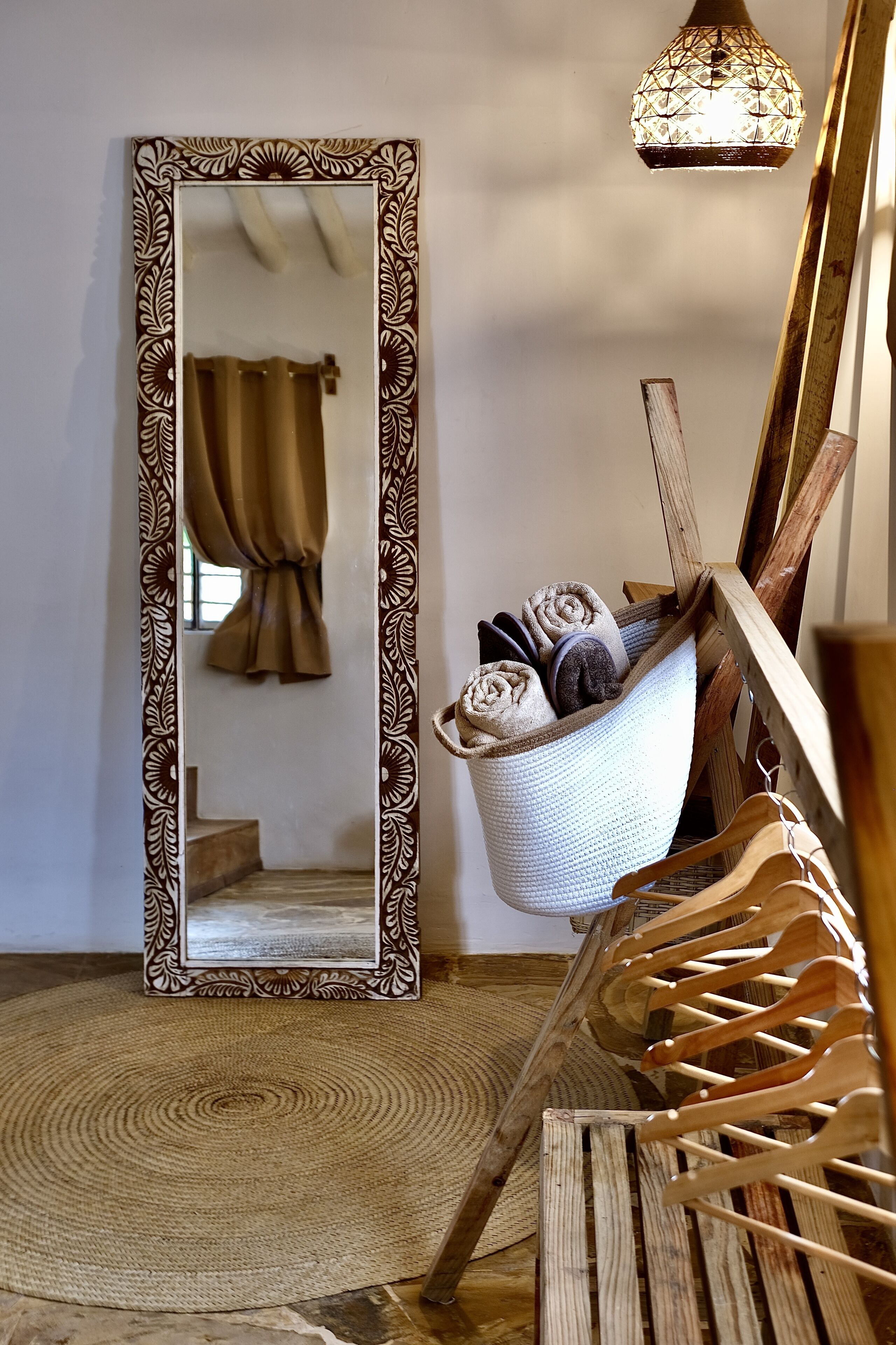 A mirror with a decorative frame, a hanging light fixture, a wooden chair with a woven seat, and a basket with rolled towels.