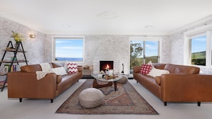 A spacious living room with two leather sofas, a glass coffee table, and a fireplace. Large windows offer views of the outdoors. - Halcyon– Breathtaking ocean views (Kiama Heights)