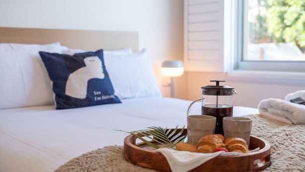 A neatly made bed with white linens, a blue pillow with a silhouette design, a coffee pot, two mugs, and a tray with croissants and a plant. - Halcyon– Breathtaking ocean views (Kiama Heights)