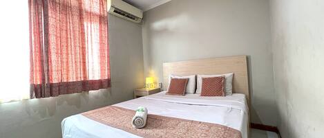 A hotel room with a bed, a nightstand, an air conditioning unit, and a window with curtains.