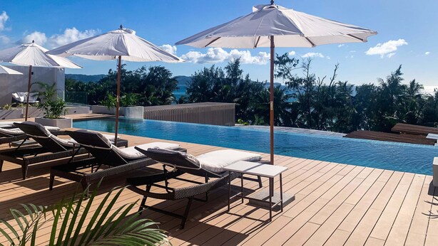 Ein Poolbereich mit Liegestühlen und Sonnenschirmen, ein Holzdeck und Blick aufs Meer.