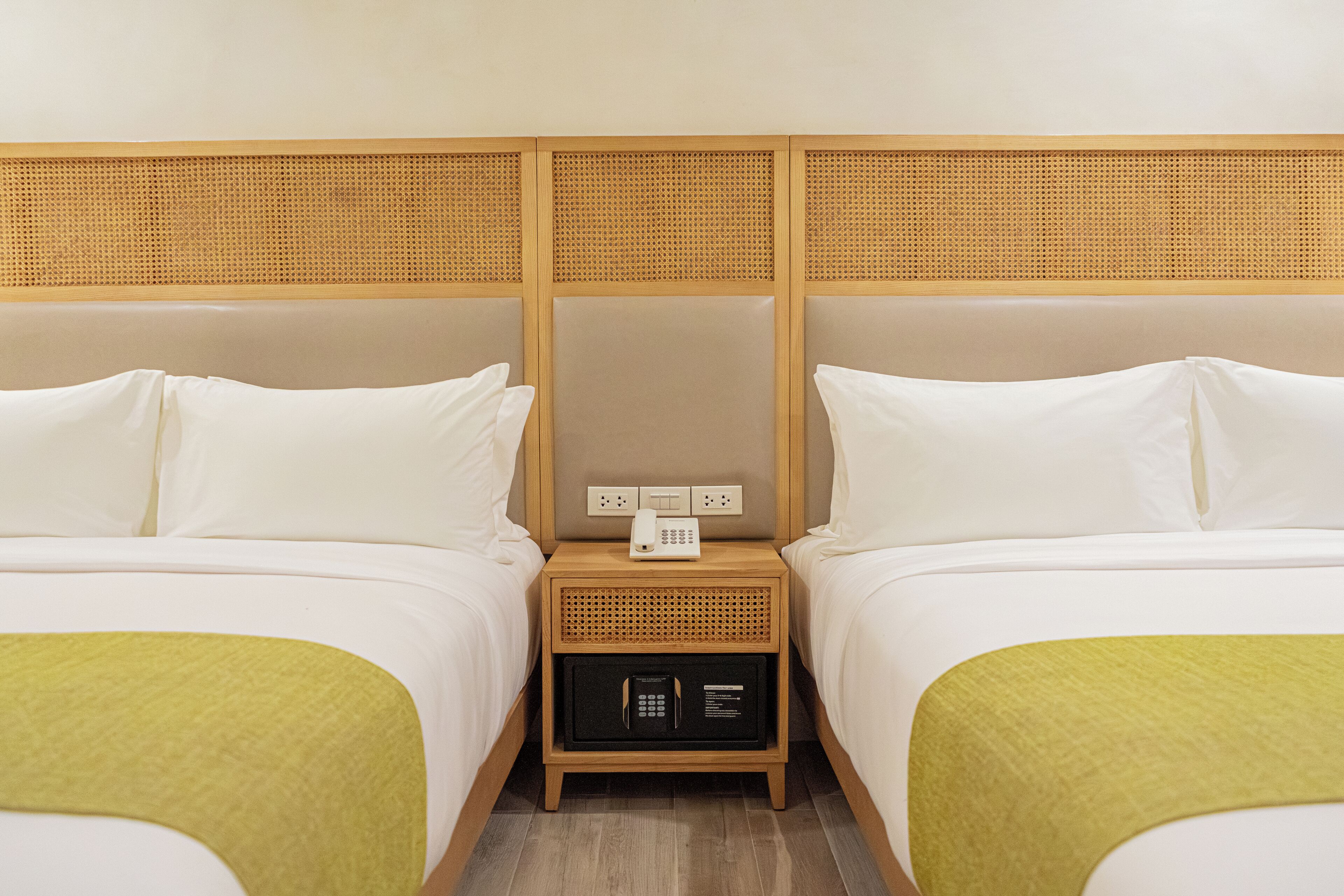 A hotel room with two beds, a nightstand with a phone and a safe, and a wooden headboard.