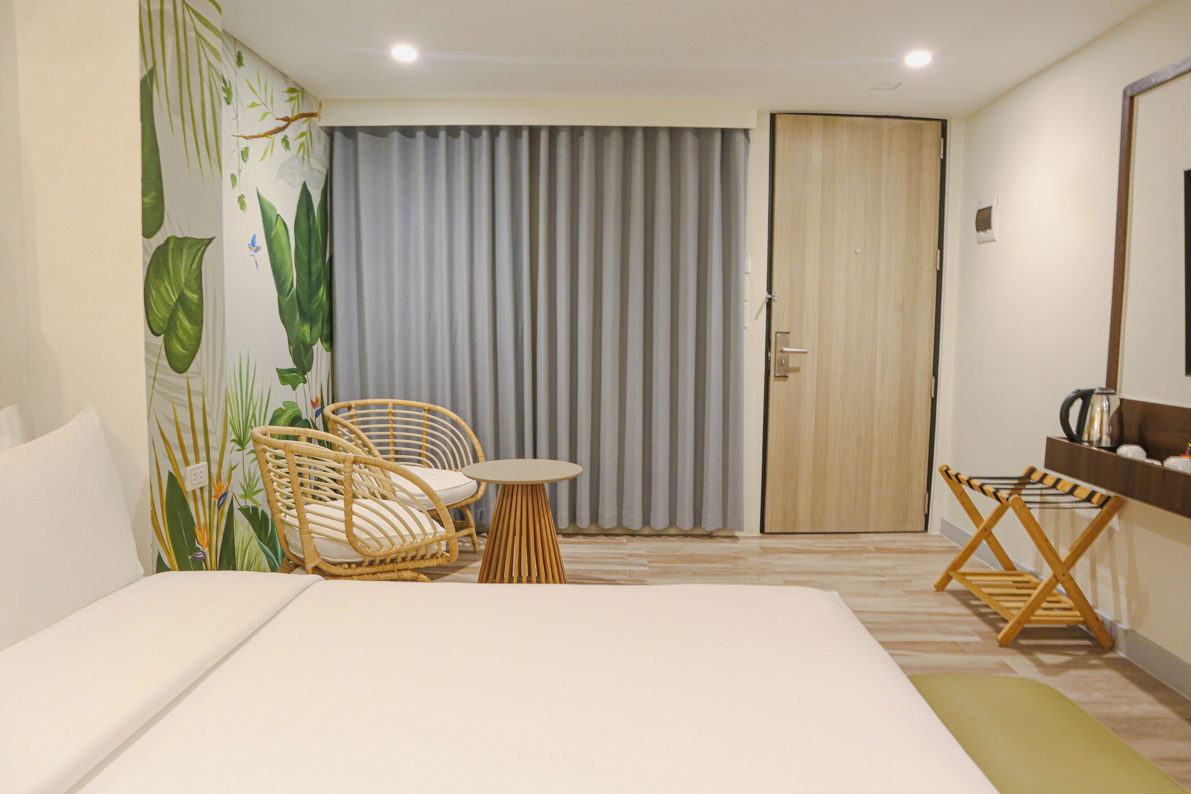 A hotel room with a bed, a wicker chair, a small round table, a wooden door, and a curtain.