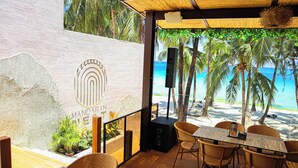 Ein Restaurant direkt am Strand mit Korbstühlen, einem Tisch mit Speisekarte und Blick auf das Meer und Palmen.