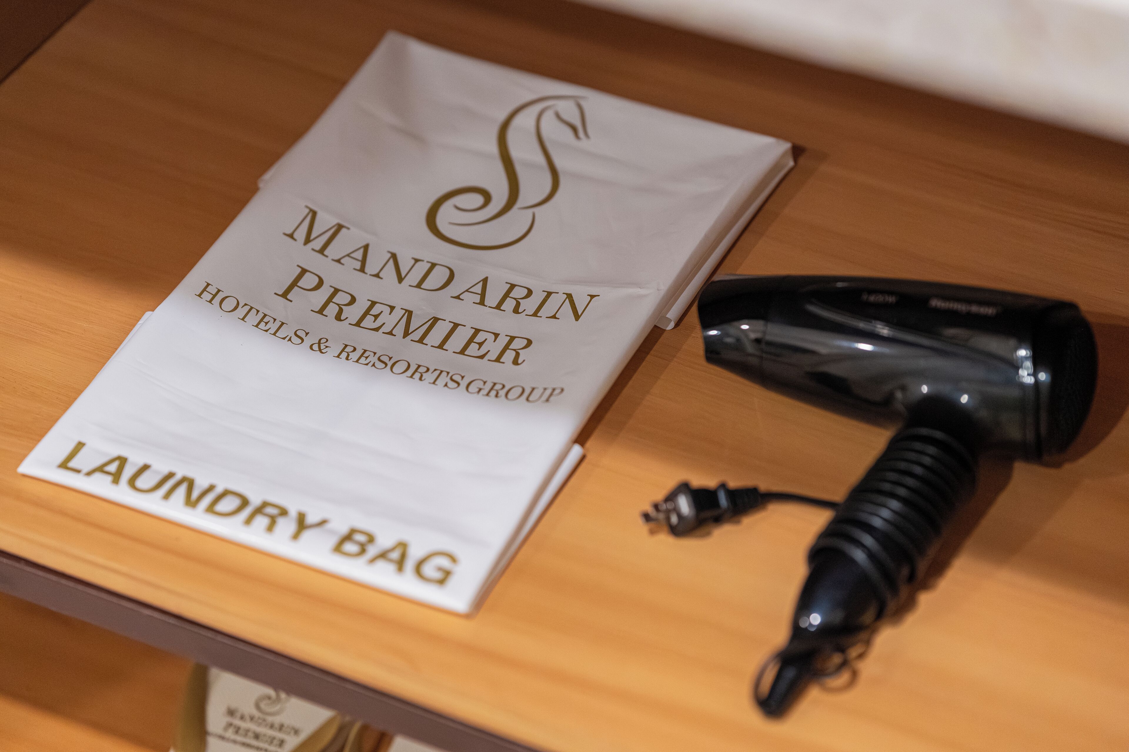 A white bag labeled "MANDARIN PREMIER HOTELS & RESORTS GROUP LAUNDRY BAG" on a wooden surface next to a black hairdryer.