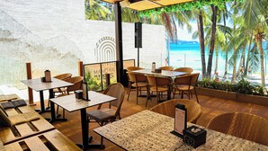 Ein Restaurant im Freien mit Holztischen und -stühlen, einem Speisekartenständer und Blick auf Strand und Palmen.