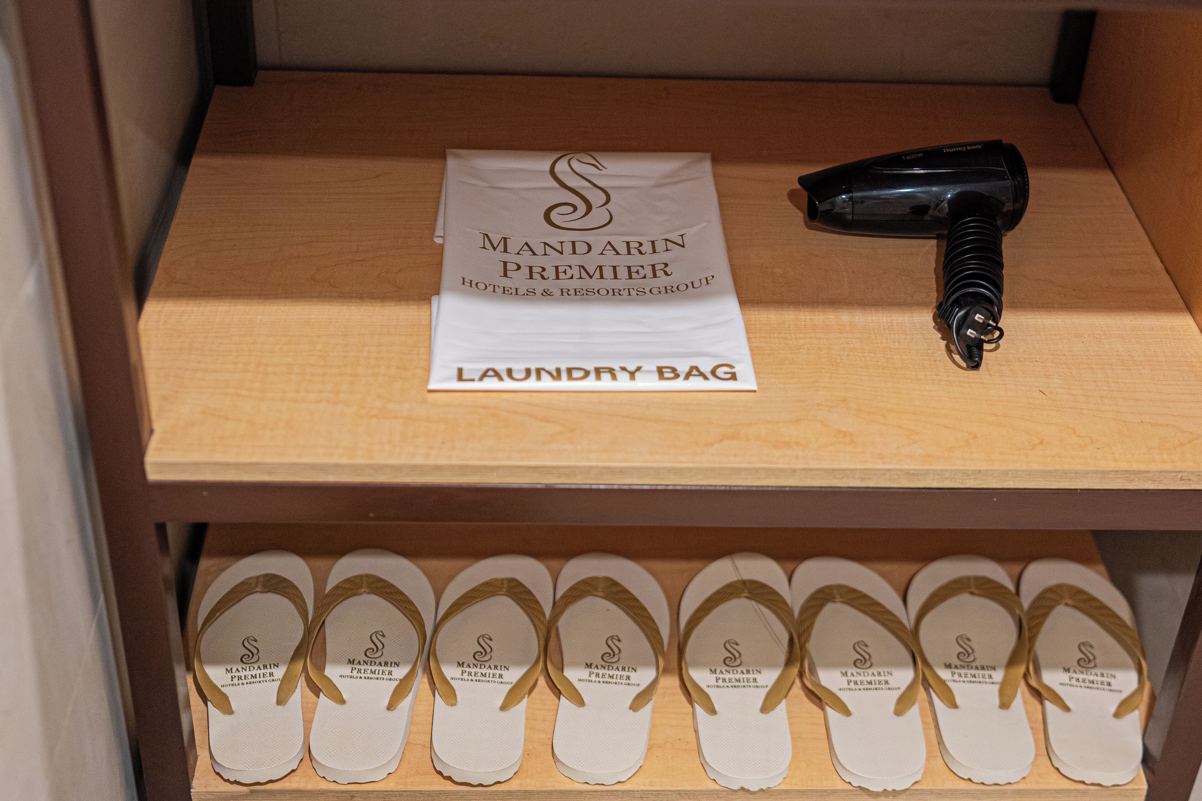 A wooden shelf with a white 'Mandarin Premier Hotels & Resorts Group' laundry bag, a black hairdryer, and multiple pairs of flip-flops with the same branding.