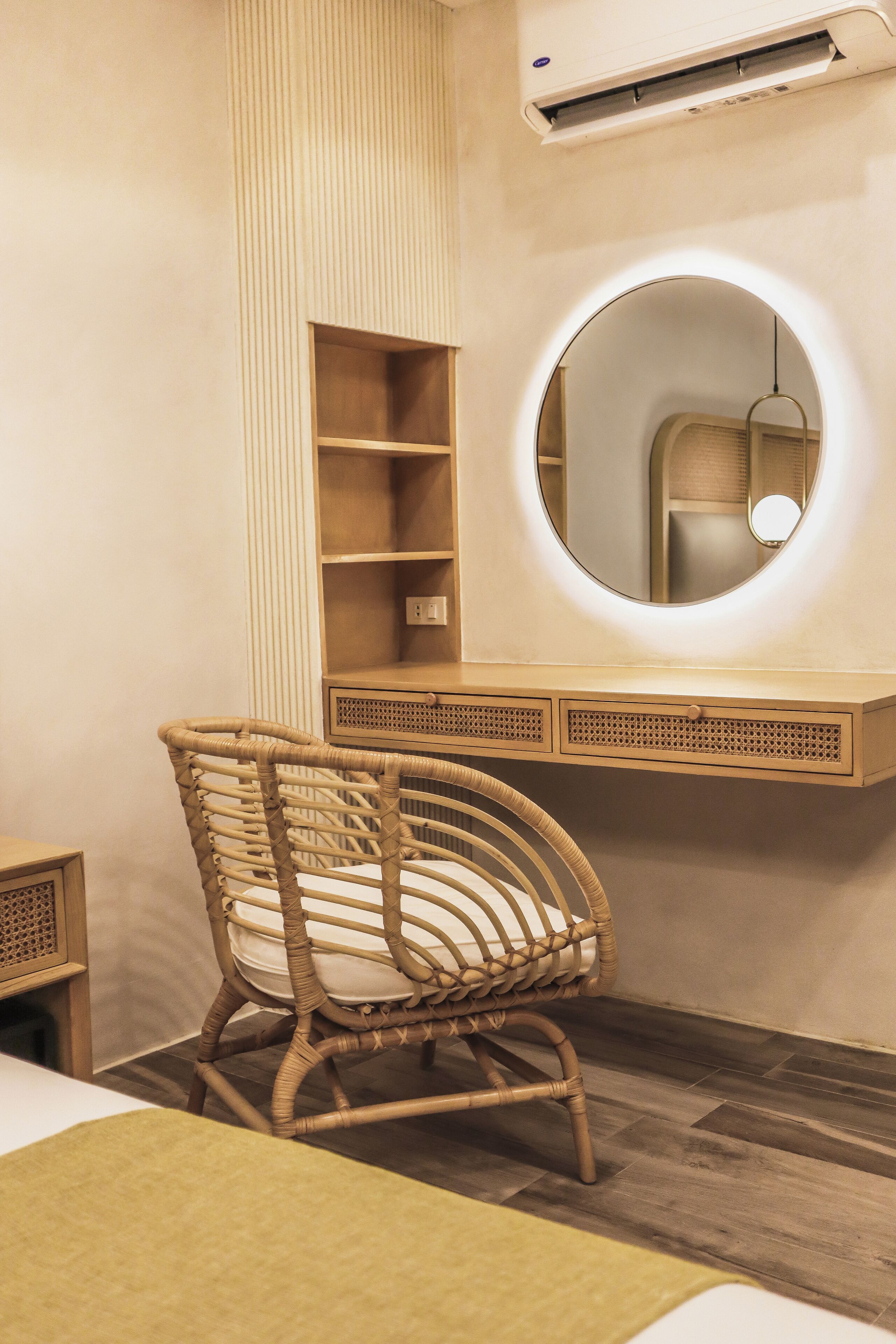 Una habitación con una silla de mimbre, un espejo redondo con luz, una estantería de madera y un equipo de aire acondicionado.