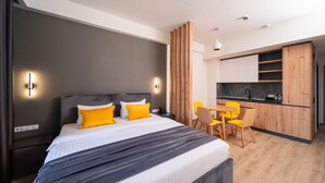 A modern hotel room with a large bed, a small kitchen area, and a dining table.