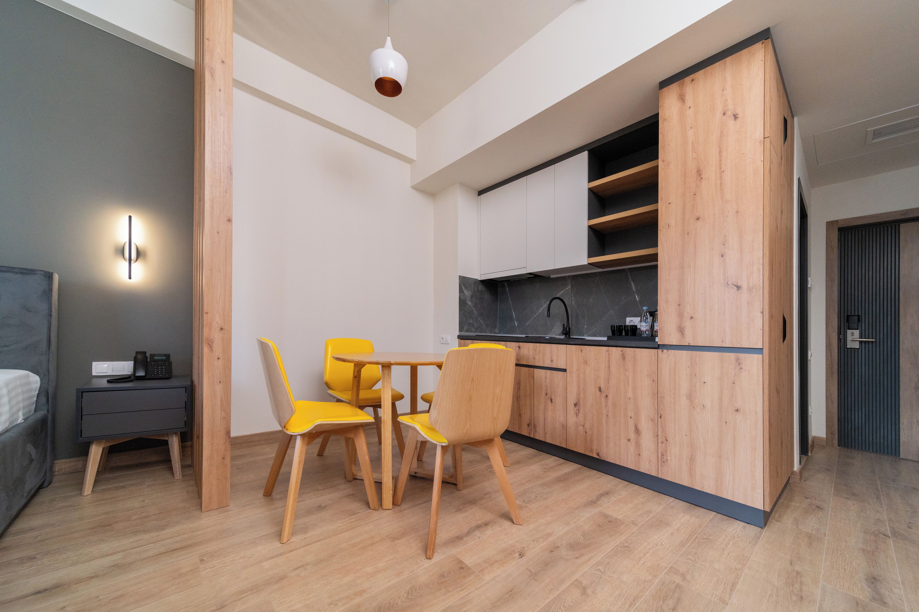 A compact living space with a kitchenette, a dining area with yellow chairs, a bed, and a nightstand with a phone.