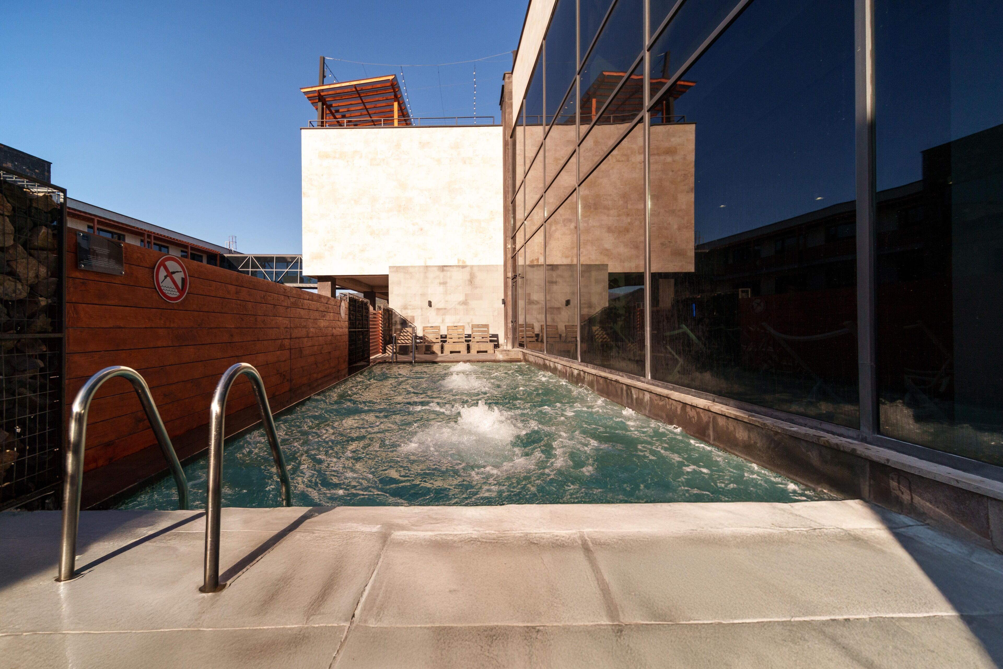 Una piscina con un cartel de prohibido bucear, rodeada por un muro de piedra y edificios modernos.
