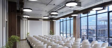 Uma sala de conferências com fileiras de cadeiras, janelas amplas e vista para uma construção externa.