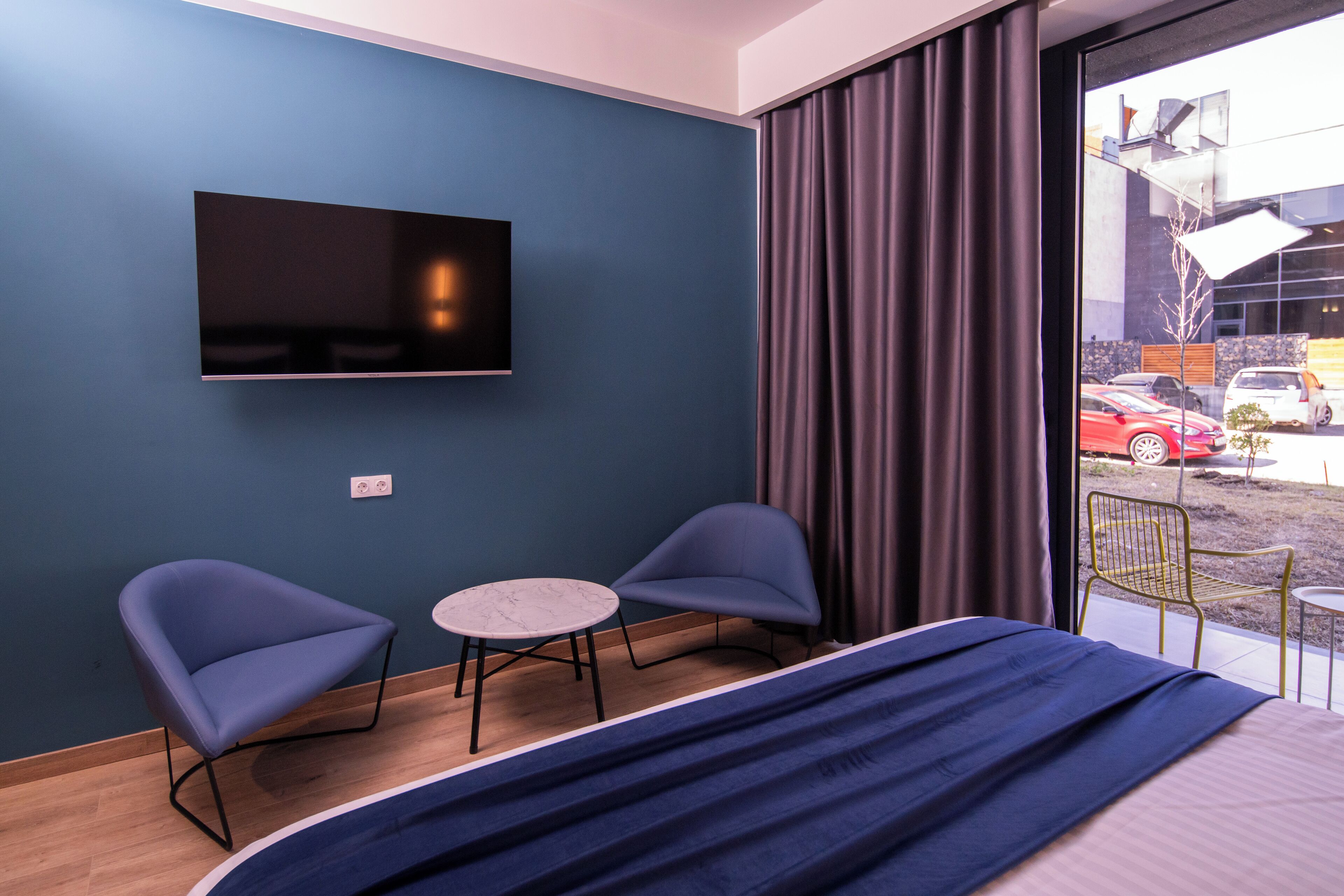Une chambre d’hôtel comprenant un lit, deux fauteuils, une petite table ronde, un téléviseur à écran plat fixé au mur et une vue sur la rue grâce à de grandes fenêtres.