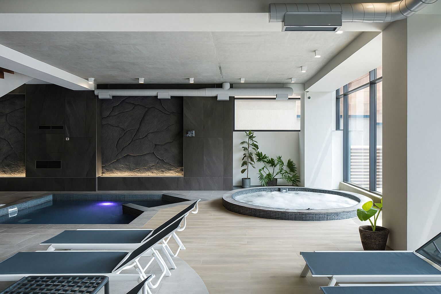 A modern indoor pool area with a rectangular and a circular hot tub, seating areas, and a large window providing natural light.