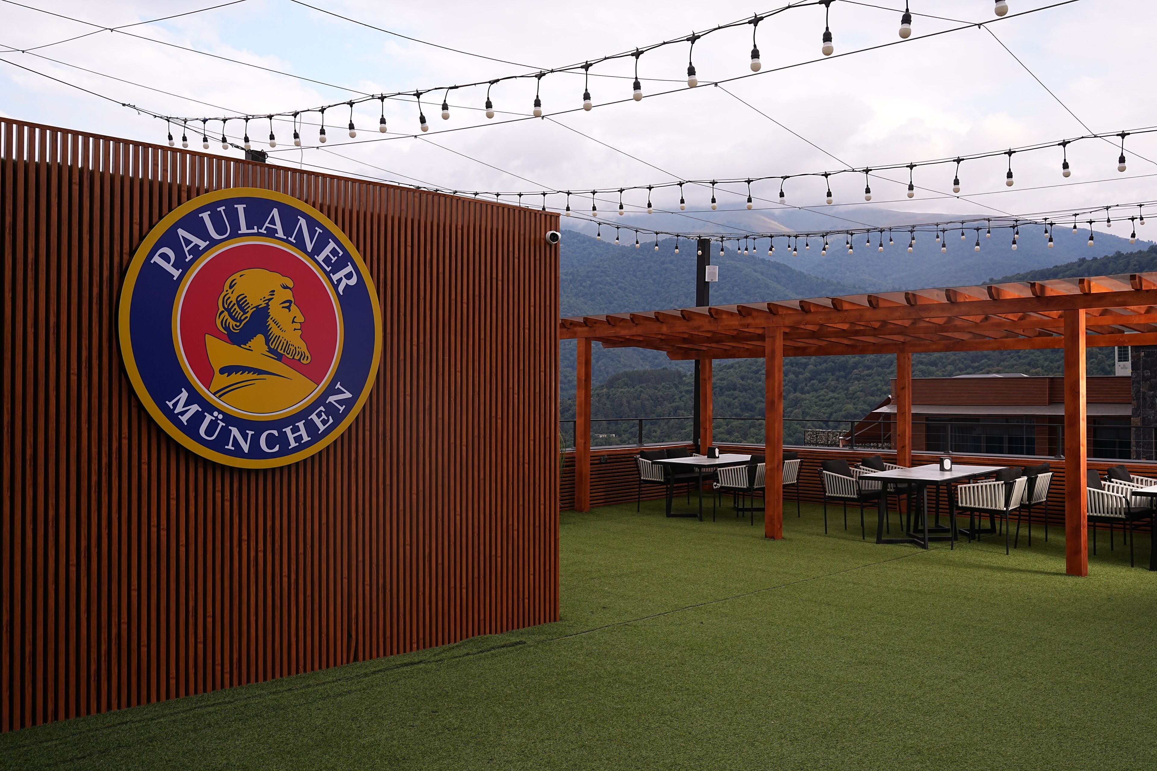 A restaurant with a sign reading "Paulaner München" and outdoor seating with a view of mountains.