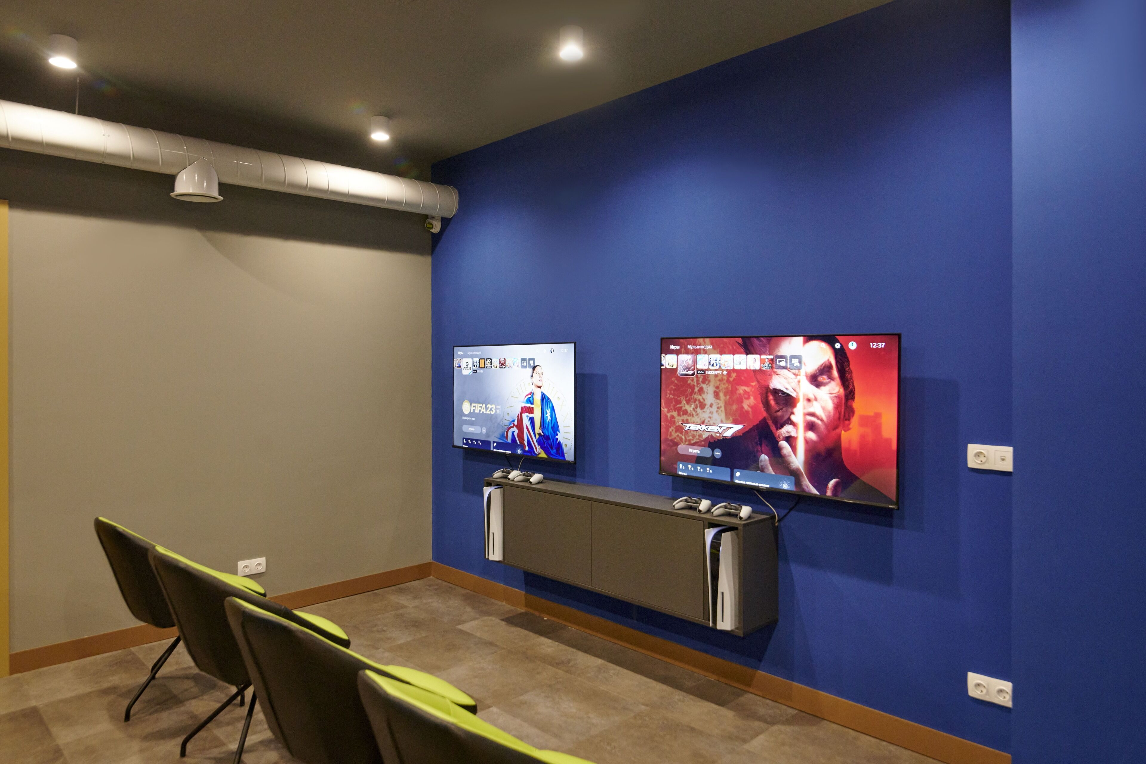 Une pièce équipée de deux téléviseurs fixés au mur, diffusant des programmes sportifs et des films, d’une console et d’un coin salon avec des fauteuils.