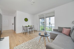 Een moderne woonkamer met een grijze bank, een eettafel met stoelen, een groot raam met zonwering en een kamerbreed tapijt met patroon.