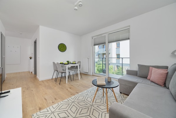A modern living room with a grey sofa, a dining table with chairs, a large window with blinds, and a patterned rug.