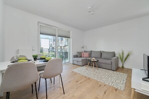 A modern living room with a grey sofa, a dining table with chairs, a TV stand, and a large window with a view of buildings. - Modern Apartment | Parking, Gym and Balcony (Wroclaw)