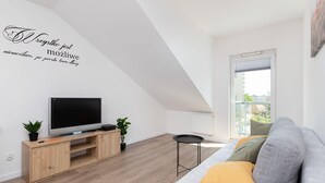 A modern living room with a flat-screen TV on a wooden TV stand, a sofa with cushions, a small round table, and a potted plant.